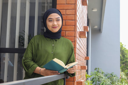 Portrait of attractive Asian hijab woman holding book in balcony. Muslim girl reading a book. Concept of literacy and knowledgeの写真素材