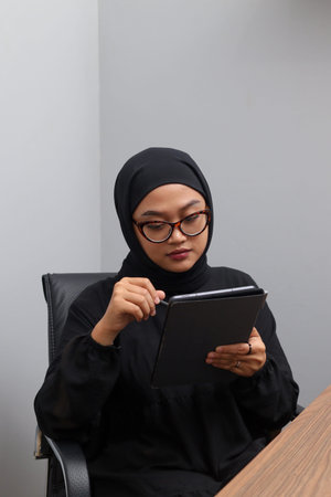 Portrait of attractive Asian hijab woman working on her tablet. Muslim girl doing task in office....