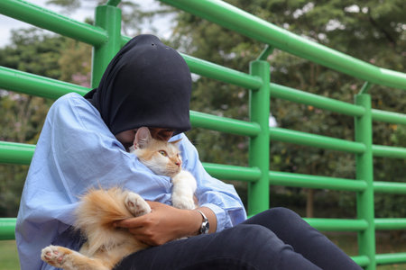 Outdoor portrait of Asian hijab woman holding and giving gentle touch to cat, taking care of her pet in nature park. Love relationship between humans and animals.の写真素材