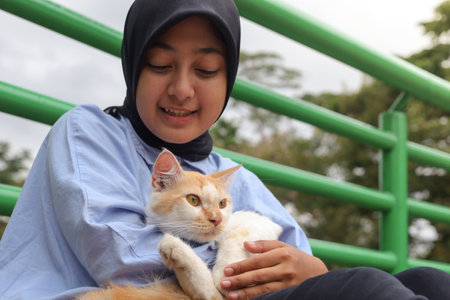 Outdoor portrait of Asian hijab woman holding and giving gentle touch to cat, taking care of her pet in nature park. Love relationship between humans and animals.の写真素材