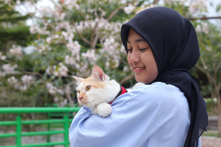 Outdoor portrait of Asian hijab woman holding and giving gentle touch to cat, taking care of her pet in nature park. Love relationship between humans and animals.の写真素材