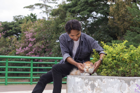 Outdoor portrait of Asian man holding and giving gentle touch to cat, taking care of his pet in nature park. Love relationship between humans and animals.の写真素材