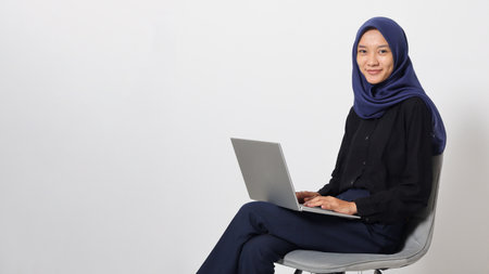 Portrait of excited Asian hijab woman in casual shirt sitting on chair, working on laptop. An employee or student doing her task and job . Businesswoman concept. Isolated image on white backgroundの写真素材