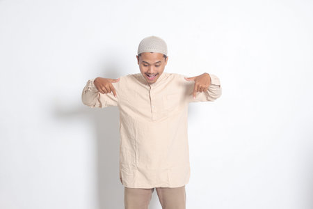 Portrait of shocked Asian muslim man in koko shirt with skullcap showing product and pointing with...