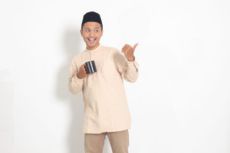 Portrait of attractive Asian muslim man in koko shirt with peci holding a mug, drinking water,...
