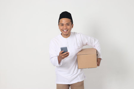Portrait of excited Asian muslim man carrying cardboard box while holding mobile phone showing cheerful expression. Going home for Eid Mubarak. Isolated image on white backgroundの写真素材