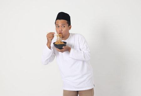 Portrait of attractive Asian muslim man eating tasteful instant noodles with chopsticks served on bowl. Iftar and pre dawn meal concept. Isolated image on white backgroundの写真素材