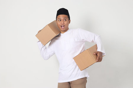 Portrait of attractive Asian muslim man in koko shirt with peci feeling grateful and peacful while carrying cardboard box. Going home for Eid Mubarak. Isolated image on white backgroundの写真素材