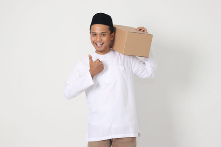 Portrait of excited Asian muslim man in koko shirt with peci carrying cardboard box while pointing and showing thumb up hand gesture. Going home for Eid Mubarak. Isolated image on white backgroundの写真素材