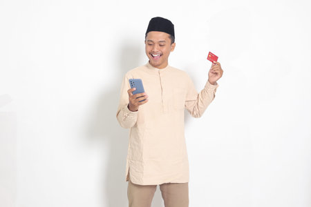 Portrait of attractive Asian muslim man in koko shirt with skullcap holding a mobile phone and...