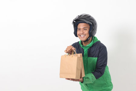 Portrait of Asian online courier driver wearing green jacket and helmet giving craft paper shopping bag with gift inside. Isolated image on gray backgroundの写真素材