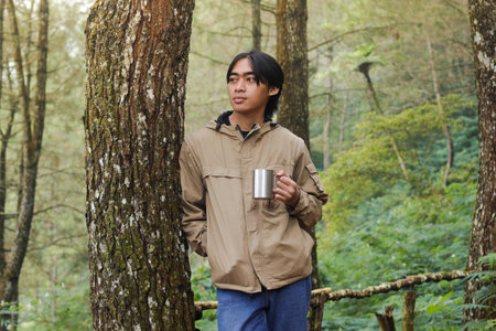 Portrait of excited Asian hiker man drinking water from stainless steel cup while trekking in nature...