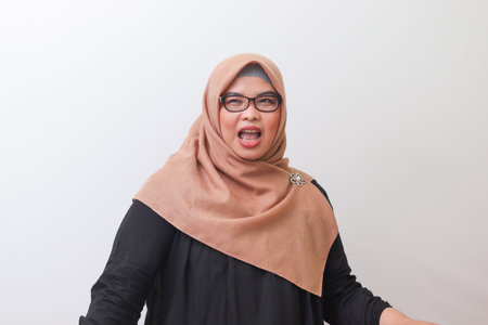 Portrait of shocked Asian woman with hijab standing against white background, laughing loudly.