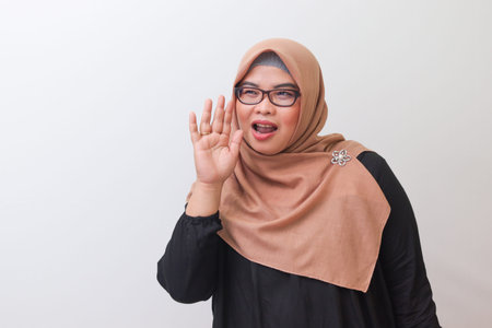 Portrait of cheerful Asian woman with hijab, holding hands near mouth and telling a secret....
