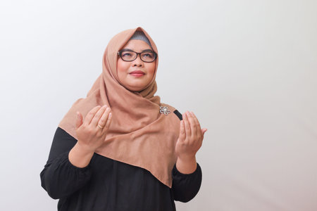 Portrait of serious Asian woman with hijab praying to God with raised hands. Isolated image on white...