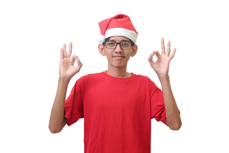 Portrait of excited Asian man in red t-shirt with santa hat doing ok approval symbol with fingers and smiling looking at camera. Isolated image on white backgroundの写真素材