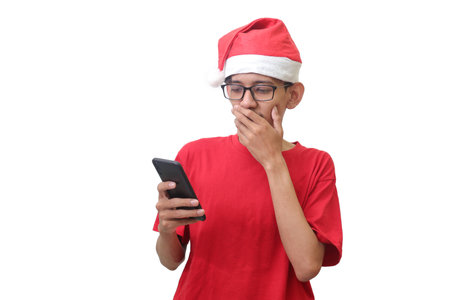Portrait of excited Asian man in red t-shirt with santa hat looking at his mobile phone while covering his mouth with fingers. Wow face expression. Isolated image on white backgroundの写真素材