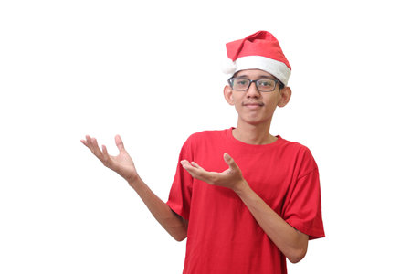 Portrait of excited Asian man in red t-shirt with santa hat, showing product, pointing up at something and smiling with the other hand on hip. Advertising concept. Isolated image on white backgroundの写真素材