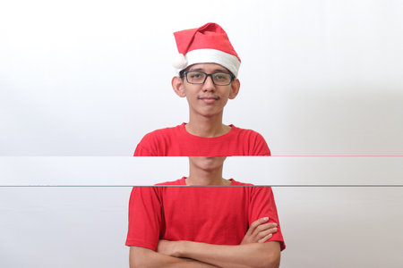 Portrait of excited Asian man in red t-shirt with santa hat keeping arms crossed and looking at camera. Isolated image on gray backgroundの写真素材