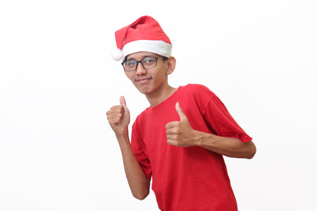 Portrait of excited Asian man in red t-shirt with santa hat smiling and looking at camera, making thumbs up hand gesture. Isolated image on gray backgroundの写真素材