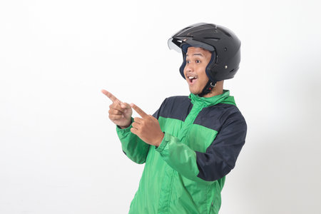 Portrait of Asian online taxi driver wearing green jacket and helmet pointing at empty space with finger. Isolated image on white backgroundの写真素材