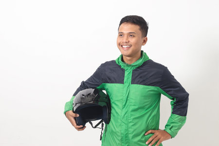 Portrait of Asian online taxi driver wearing green standing against white background, smiling and looking at camera, holding his helmet on the sideの写真素材