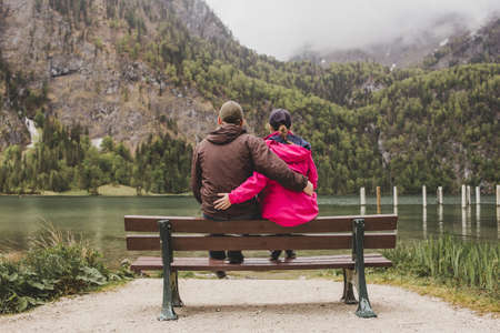 Lovely couple sit on the bench & embrace on background of alpine lake Konigssee, rock mountain & cloud sky. Bavaria. Germanyの写真素材