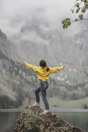 Woman in yellow sweatshirt feels freedom on the stone on background of Obersee lake, Rothbach waterfall, rock mountain & green forest in cloud weather. Bavaria. Germany. Vertical wallpaperの写真素材