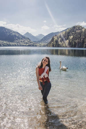 Brunette caucasian woman stands knee-deep in the water near white swan & smiles on background of mountains & pine forest & blue sky in Bavaria. Alpsee. Germanyの写真素材
