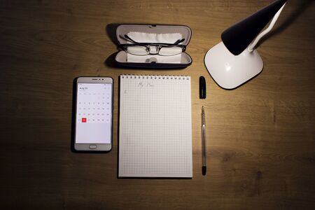 Education concept - notebook on a wooden table with hand written word plan, vision glasses in a case, pen and mobile phone with an open electronic calendar and a white lamp on top.の写真素材