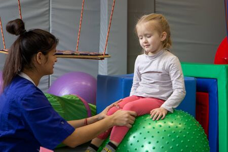 little blonde girl sitting on big gym ball working with physiotherapistの写真素材