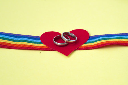 pair of white golden wedding rings on LGBTQ rainbow ribbon with red heart on yellow backgroundの写真素材