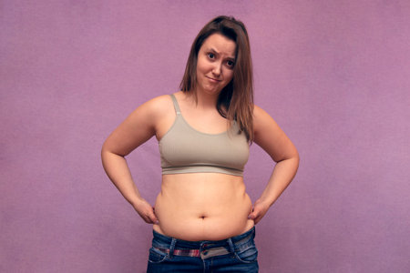 Overweight sad woman hand pinching excessive belly fat on pink background, unhealthy concept