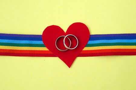 pair of white golden wedding rings on LGBTQ rainbow ribbon with red heart on yellow backgroundの写真素材