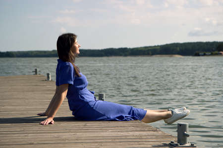 Side view of dreaming girl sitting on jetty forest and river on background. serene calm woman resting on the shore of the lake, enjoying the sun and good weatherの写真素材