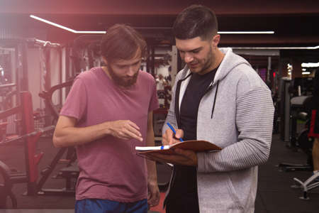 fitness, sport, exercising and diet concept - smiling young sportsman and personal trainer with clipboard writing exercise plan in gymの写真素材
