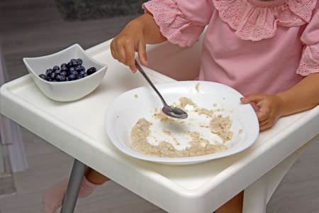 Little kid girl eating her morning porridge oatmeal with blueberries herself, sitting in baby chair in kitchenの写真素材