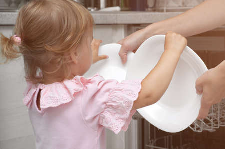 Adorable blonde toddler girl helping in the kitchen taking plates out of dish washing machine. kid helps to do household choresの写真素材