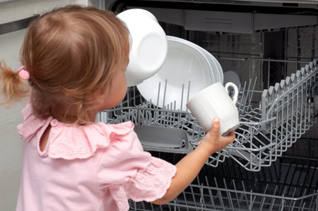 Adorable blonde toddler girl helping in the kitchen taking plates out of dish washing machine. kid helps to do household choresの写真素材