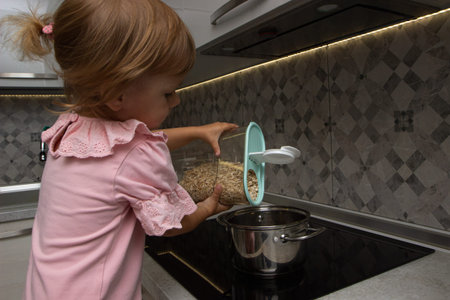preparation of a family breakfast. kid girl help cook porridge in morning. little girl pours oat...