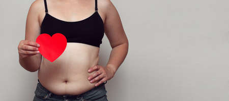 Overweight fat woman showing her belly fat with paper heart close up, selective focus. Bodysitive conceptの写真素材