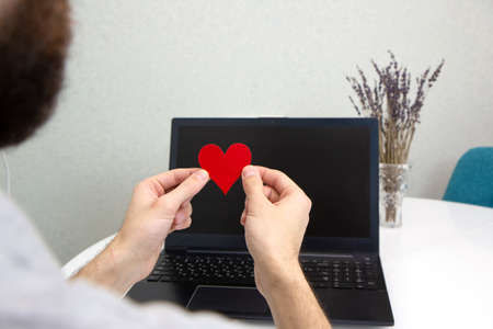 Hand holding red Valentine heart on laptop background Close-up. Online dating, love and long distance relationship concept. romantic virtual dateの写真素材