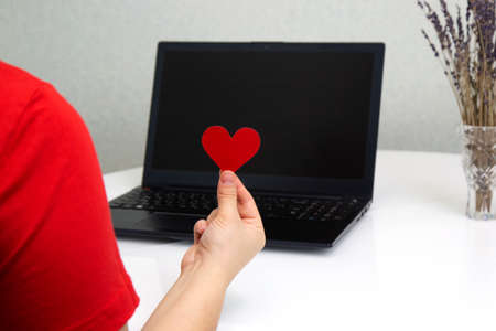 Hand holding red Valentine heart on laptop background Close-up. Online dating, love and long distance relationship concept. romantic virtual dateの写真素材