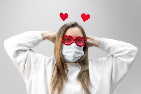 Portrait of woman in funny glasses and medical protective wearing mask holding in hands red hearts. concept of valentines day during pandemic period, valentine in the distanceの写真素材