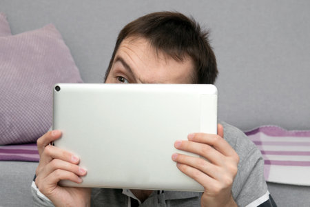 a man uses a tablet at home, looks out suspiciously from behind the screen, persecution mania and fear of surveillanceの写真素材