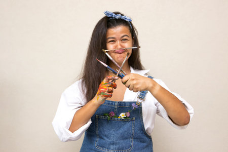 Delighted inspired artist posing in studio, holding paintbrushes looking cheerful . creative...