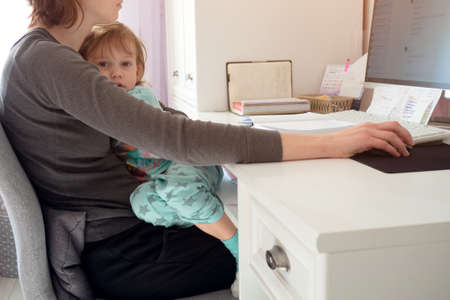 Mother holding girl kid and using computer at home with selective focus. Candid authentic and real life mom working and parenting. Home office work onlineの写真素材