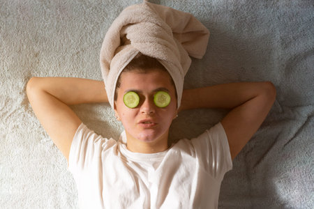 Woman with a face mask and towel on head and pieces of cucumber on her eyes. Spa and relaxing beauty...