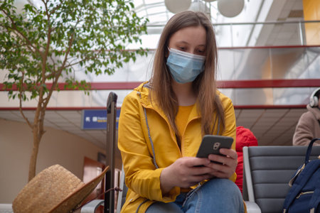Caucasian woman tourist in waiting room with medical mask protection work in smartphone. Travaler...