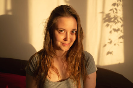 Young woman looking at camera in warm rays of sunset. Warm calm portrait of girl indoor. light and...
